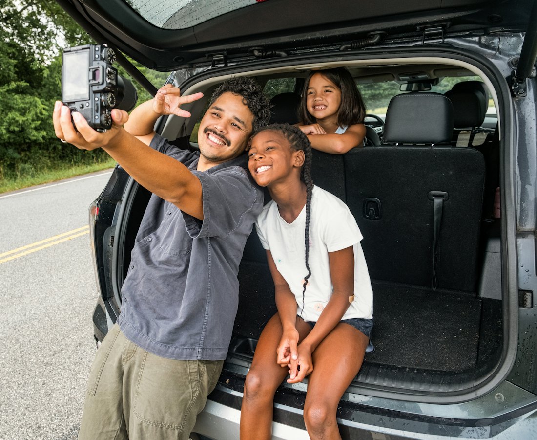 Pai e duas filhas tirando uma selfie ao lado do carro, na estrada